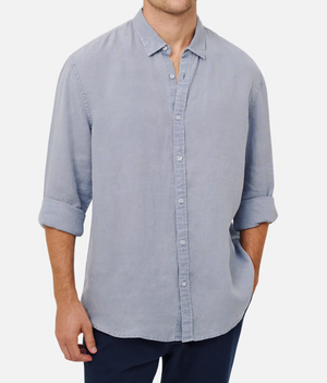 Someone stands confidently in a light blue Industrie The Tennyson Linen Shirt with rolled-up sleeves, paired with dark pants and one hand in their pocket against a white backdrop.
