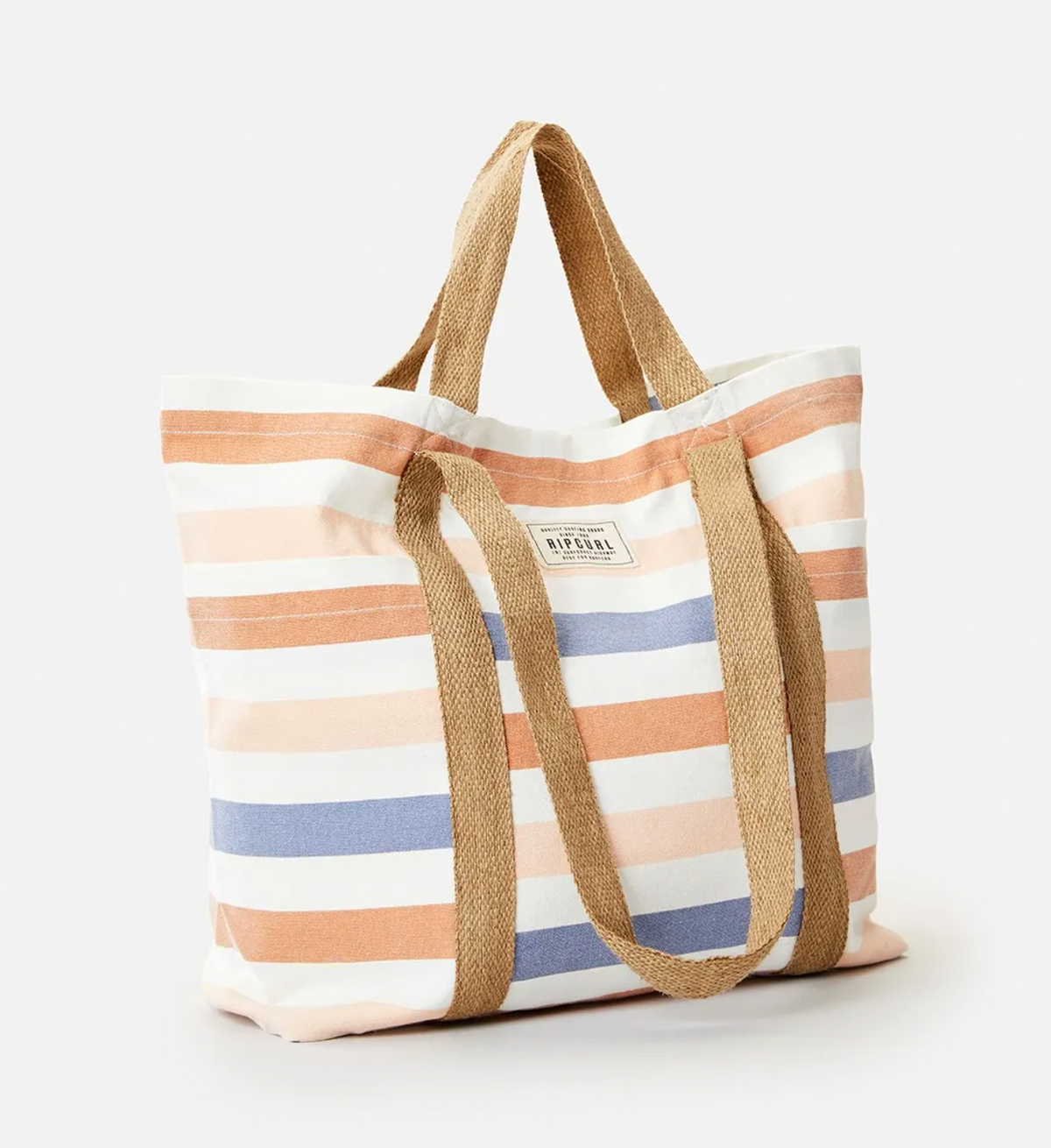The Rip Curl Organic 29L Beach Tote features beige, light blue, and orange stripes on organic cotton canvas. It includes sturdy brown handles and a small rectangular Rip Curl logo patch on the front, all set against a plain white background.