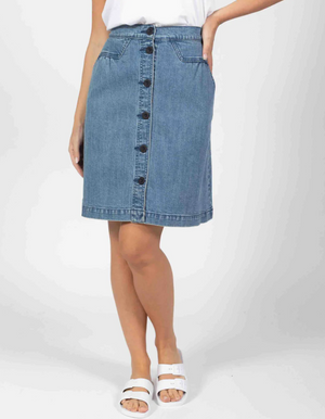 Wearing a white top and the Foxwood Amanda knee-length denim skirt featuring a button front and pockets, the person pairs this with white sandals. Their pose shows one hand resting on their hip against a plain background.