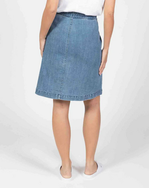 A person stands facing away, wearing the Foxwood Amanda Skirt—a knee-length blue denim skirt with an elastic waist—paired with a white top and white slip-on shoes against a plain white background.