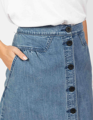 A close-up shows someone wearing the Foxwood Amanda Skirt, a high-waisted denim piece with an elastic waist and black button front. They have their hand in one of the side pockets, perfectly paired with a crisp white top.