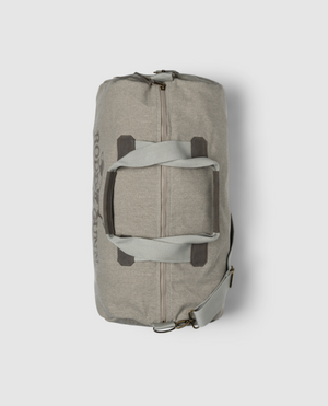 The Rodd & Gunn Double Barrel Duffle Bag is made from durable cotton canvas, featuring dark gray handles and a shoulder strap. This beige bag, with Rodd & Gunn branding, includes two brown accent straps across the middle on a light gray background.