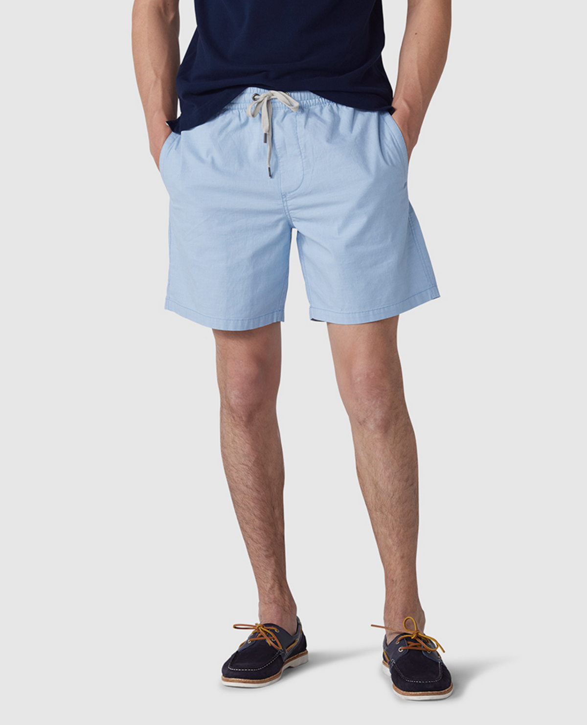 A man wears a navy-blue T-shirt, light blue Rodd & Gunn Resort Short with an elastic waist made of cotton elastane, and dark boat shoes with light brown laces. His hands are in his pockets against a plain white background.