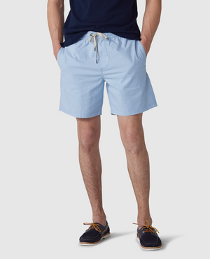 A man wears a navy-blue T-shirt, light blue Rodd & Gunn Resort Short with an elastic waist made of cotton elastane, and dark boat shoes with light brown laces. His hands are in his pockets against a plain white background.