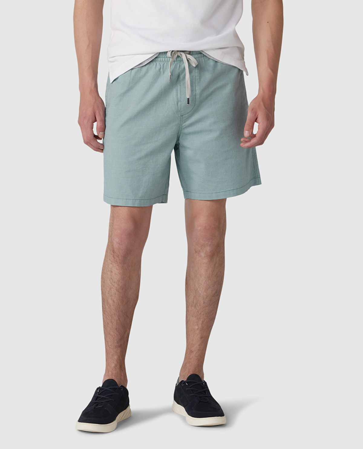 Wearing a white t-shirt, black sneakers, and Rodd & Gunn Resort Shorts in light green with an elastic waist, the individual stands against a plain background. These knee-length shorts are crafted from cotton elastane for comfort and ease of movement.