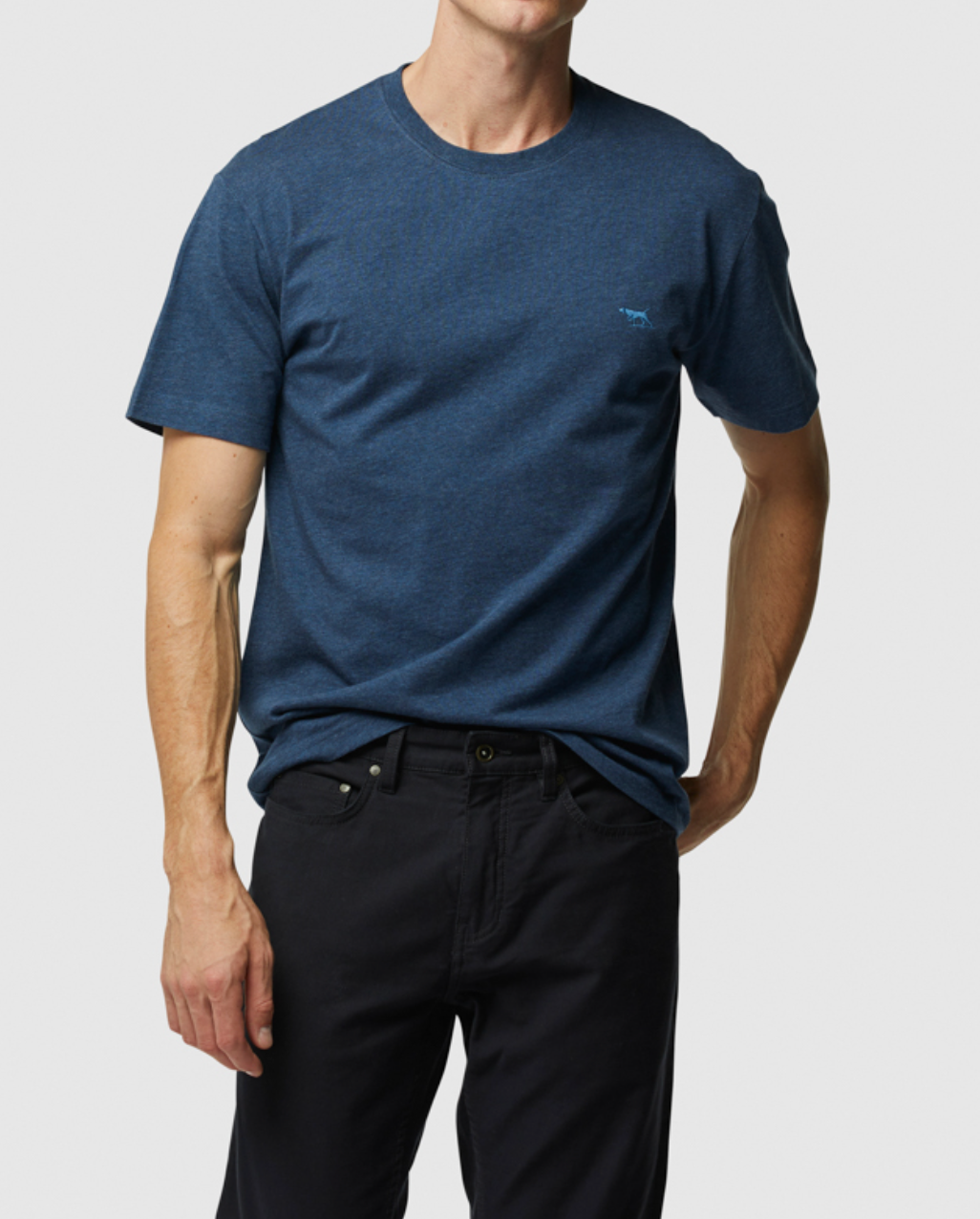 A person wearing Rodd & Gunn's dark blue 'The Gunn T-Shirt' with a subtle left chest logo and black pants stands against a plain white background, their right hand casually in their pocket.