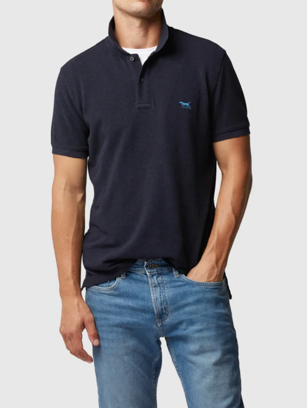 A person wears the Rodd & Gunn The Gunn Polo in dark navy with a horse logo and light blue jeans. Made from Australian Cotton, it exudes casual elegance. Their left hand is in their pocket while the right arm rests by their side against a plain light grey background.