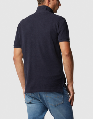 A man with short hair, wearing a Rodd & Gunn "The Gunn" Polo in dark navy made from Australian cotton and light blue jeans, stands facing away against a plain gray backdrop.