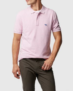 A man wears the Rodd & Gunn The Gunn Polo in light pink, a modern fit made from Australian Cotton with a small blue chest logo, paired with olive green pants, standing against a plain light background.