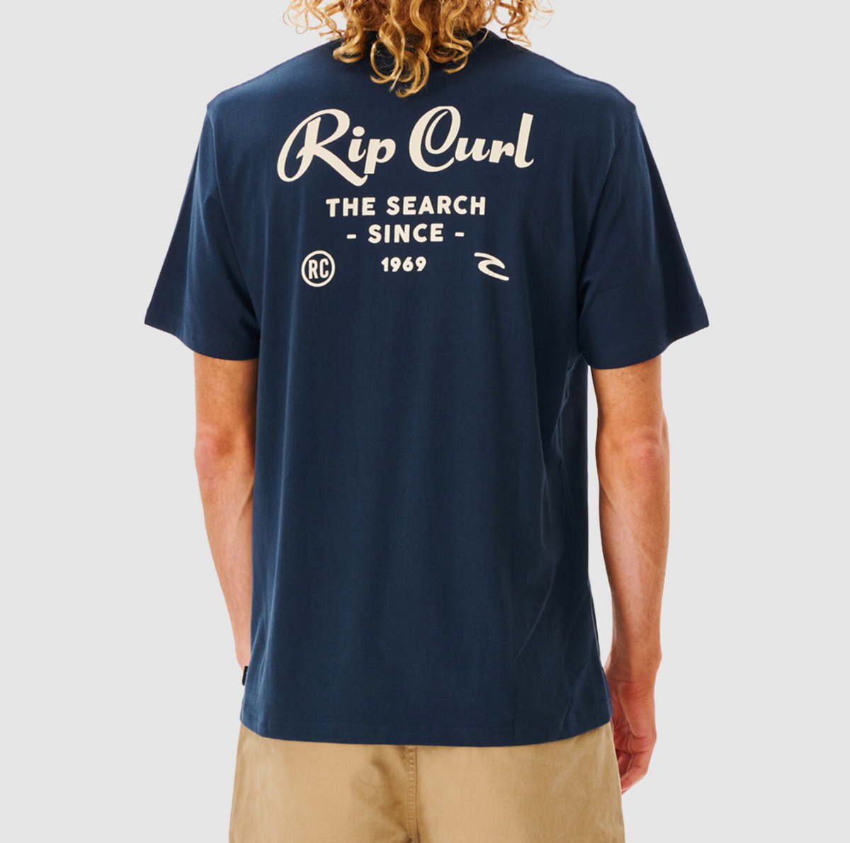 Person with curly hair wearing a comfortable navy blue Rip Curl Twist Tee, featuring white "Rip Curl The Search Since 1969" text and logos on the back, standing with hands by sides. Paired with khaki shorts against a light gray background.