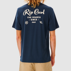 Person with curly hair wearing a comfortable navy blue Rip Curl Twist Tee, featuring white "Rip Curl The Search Since 1969" text and logos on the back, standing with hands by sides. Paired with khaki shorts against a light gray background.