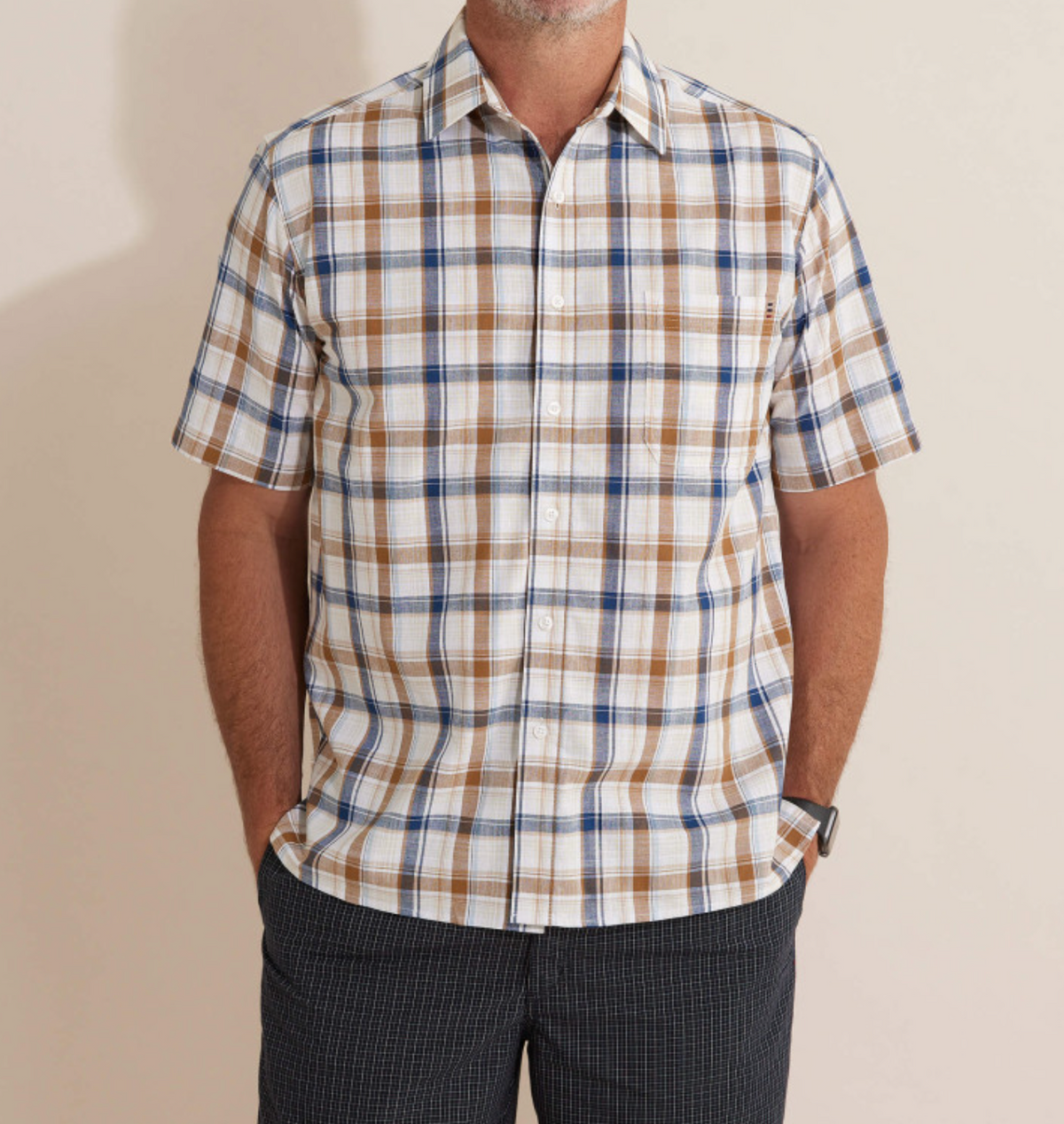 The Breakaway Amber Flaxley Shirt, featuring a comfortable short-sleeve plaid design in white, blue, and brown, complements dark checkered pants as the person rests their hands in the pockets. Their casual ensemble stands out against the plain background with their face not visible.