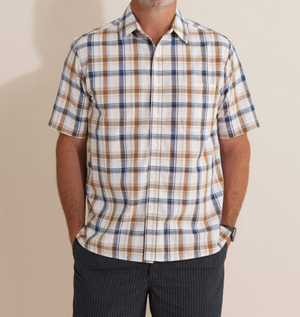 The Breakaway Amber Flaxley Shirt, featuring a comfortable short-sleeve plaid design in white, blue, and brown, complements dark checkered pants as the person rests their hands in the pockets. Their casual ensemble stands out against the plain background with their face not visible.