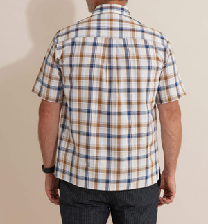 A person in a Breakaway Amber Flaxley Shirt, a comfortable short-sleeved plaid made of 100% cotton in white, brown, and blue, is seen from behind. They wear dark-textured pants and a wristwatch on their left wrist; the background is a plain light color.