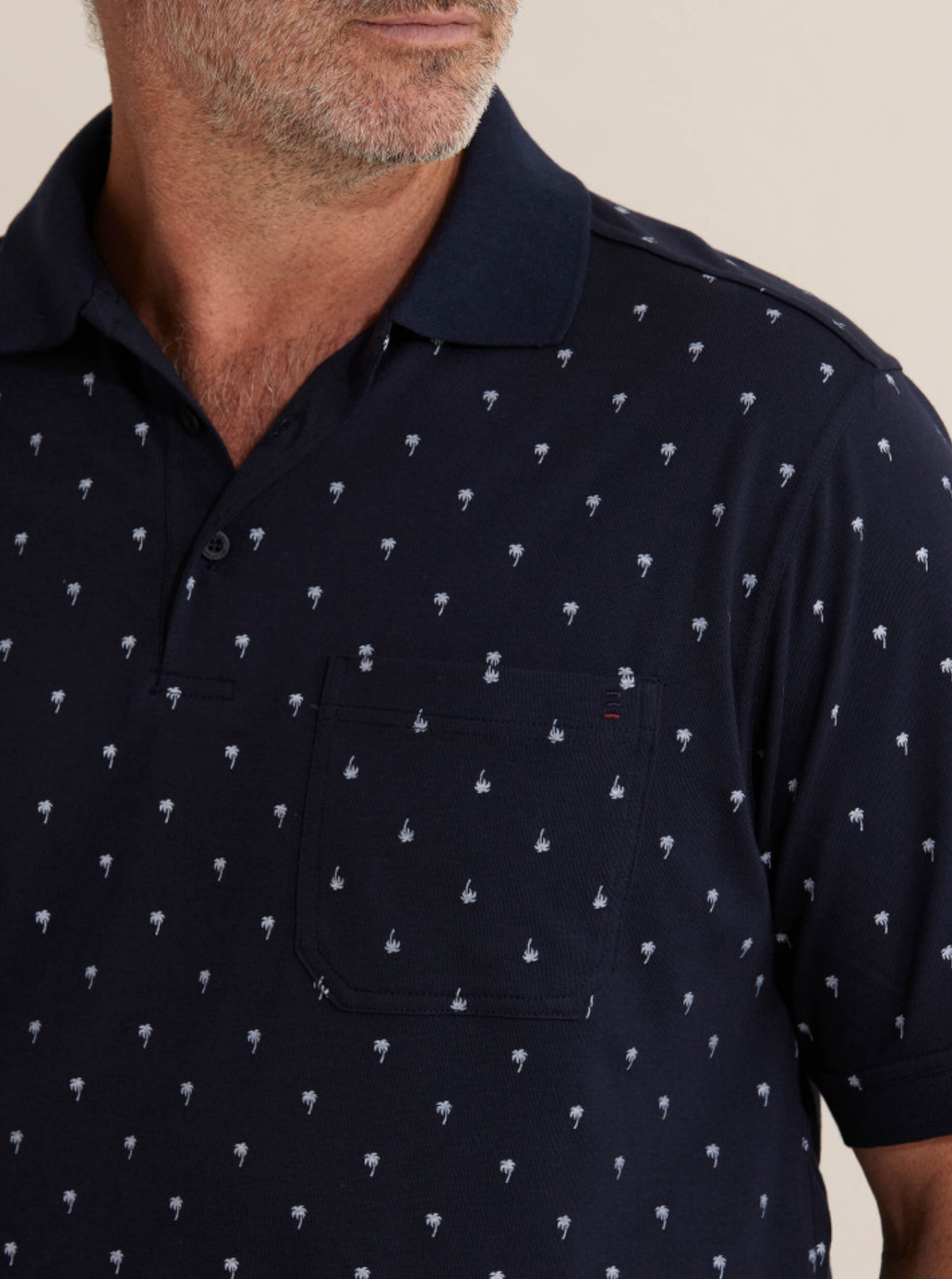 A person is wearing the Breakaway Urban Polo, a comfortable navy blue shirt adorned with small white palm tree patterns. It features a chest pocket and collar, complementing their facial hair and short hair styled in a way that hints at a breakaway style with the head slightly turned to one side.