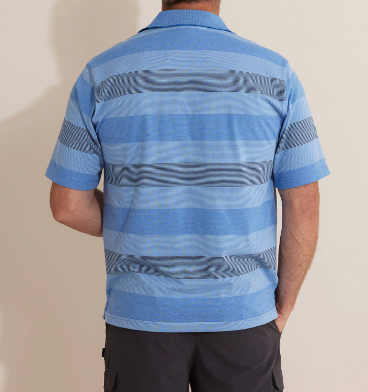 A man is seen from behind wearing a Breakaway Retba Kinnersly Polo with blue and grey stripes, short sleeves, and a rib knit collar. His left hand is in the pocket of black shorts against a plain light background.