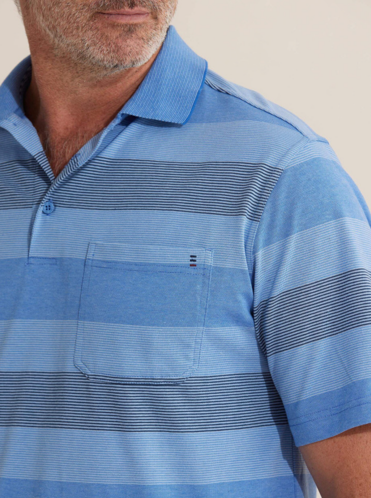 A man stands against a plain background wearing the Breakaway Retba Kinnersly Polo, featuring blue horizontal stripes, short sleeves, a rib knit collar, and a chest pocket. His face is partially visible in the scene.