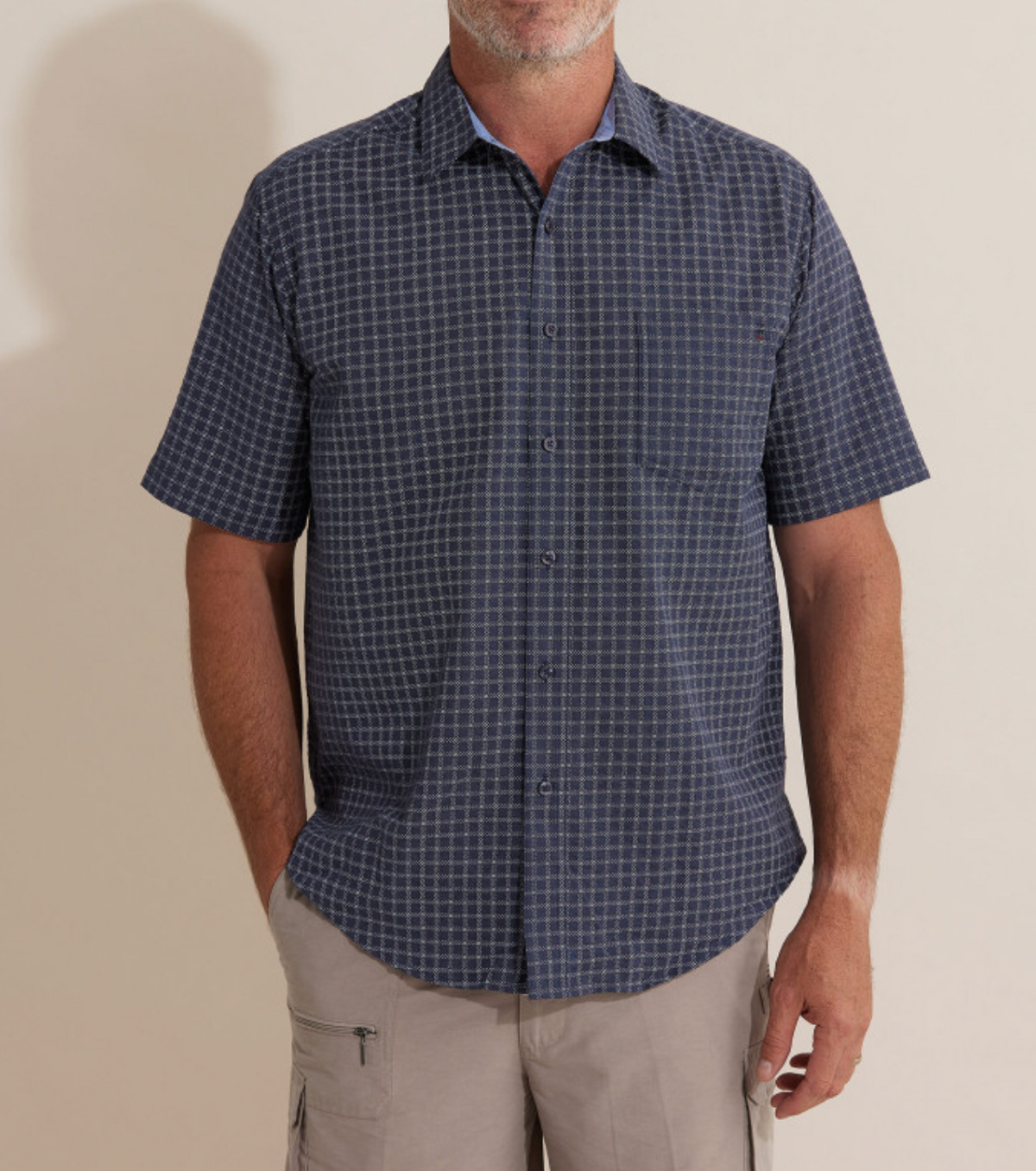 A man stands against a neutral background wearing the Breakaway Skye Seersucker Shirt, featuring short sleeves and a navy blue checkered design. He has beige cargo shorts on and rests one hand in his pocket. The image is cropped from the neck down.