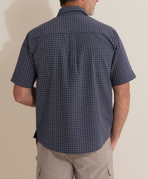 A man with short dark hair and a beard faces away, styling in a Breakaway Skye Seersucker Shirt, a navy short-sleeved checkered shirt made from 100% cotton, paired with light beige shorts. He stands against a plain, light-colored background, showcasing casual breakaway style.