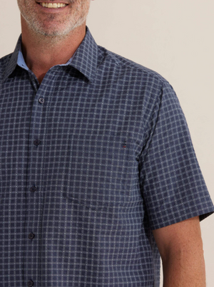 A man is wearing the Breakaway Skye Seersucker Shirt, a short-sleeved, blue patterned button-up made of 100% cotton. The photo captures his shoulders and upper arms, with a slight smile and beard against a plain background.