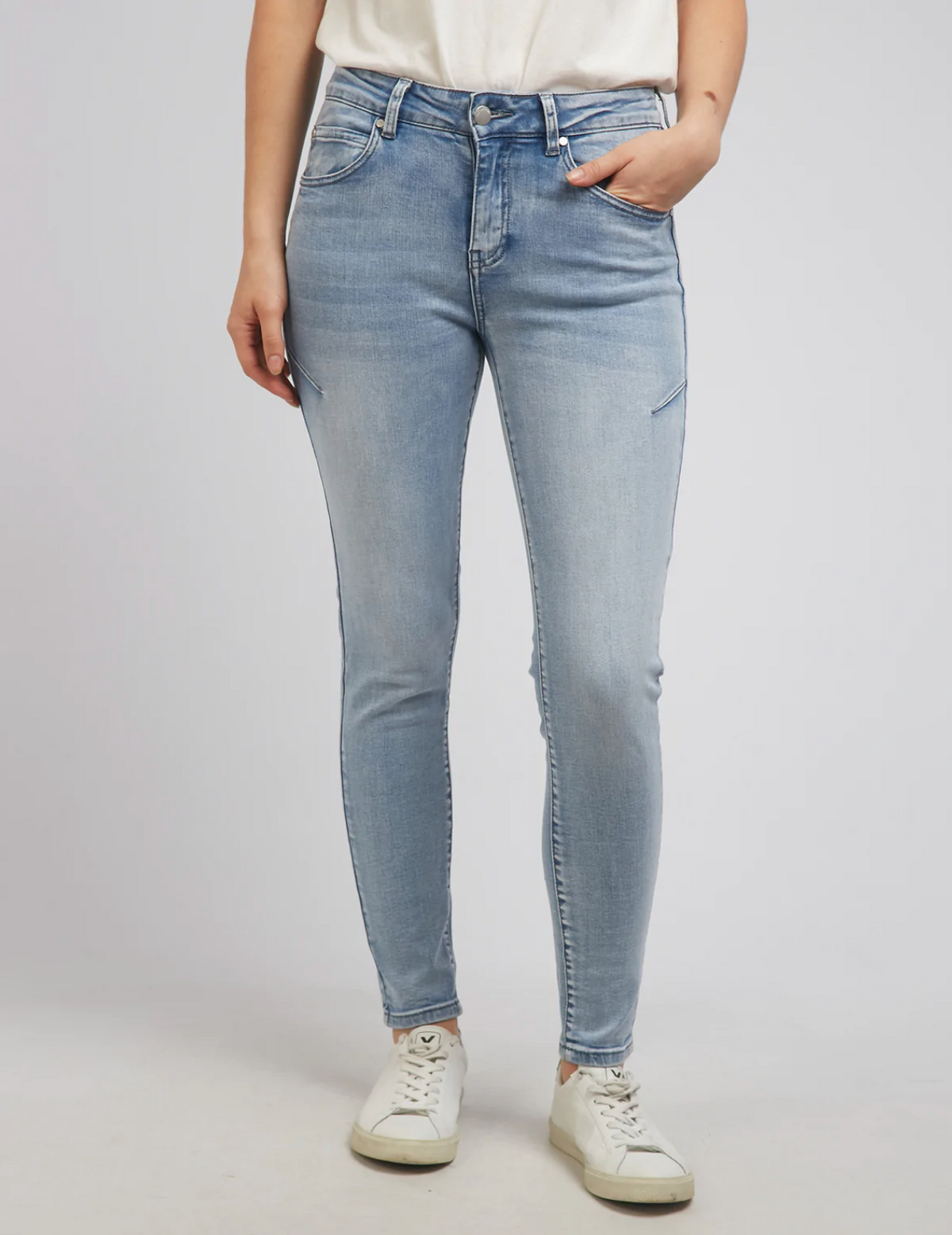 A person wears Foxwood City Jeans in light blue skinny fit, mid-rise waist, paired with a white T-shirt and crisp white sneakers, standing with one hand in their pocket against a plain light gray background.