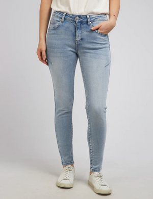 A person wears Foxwood City Jeans in light blue skinny fit, mid-rise waist, paired with a white T-shirt and crisp white sneakers, standing with one hand in their pocket against a plain light gray background.