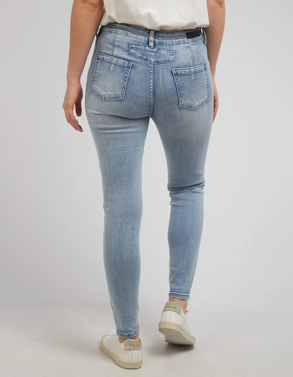 Rear view of a person in Foxwood City Jean with a mid-rise waist and skinny leg fit, paired with white sneakers. Set against a plain background, the jeans showcase visible back pockets and subtle patch detailing. A crisp white top completes the look.