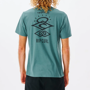 A person with curly hair wears a casual, organic cotton Rip Curl Search Icon Tee in teal, featuring black graphic designs of stylized waves and the words "The Search" and "Rip Curl" on the back. They pair it with black shorts against a plain white background.