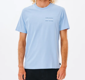 The individual wears a light blue Rip Curl Revival Repeater Tee made from 100% Cotton Jersey with subtle text on the left chest. They're paired with black pants and stand against a white background, arms at their sides, complementing their curly blond hair.