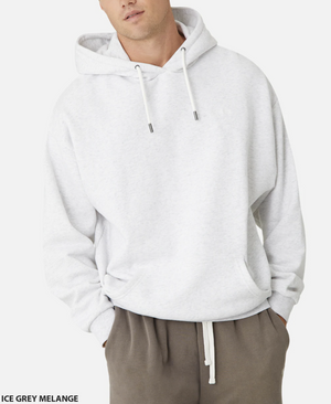 Someone wears an oversized ice grey melange Industrie The Del Sur Hoodie with the hood down, drawstrings, and the embroidered logo. Their hands are casually tucked in the pockets, and they pair it with brown pants against a plain white background.