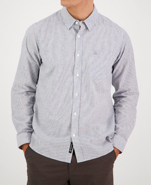A person models the Swanndri Carillo Beach L/S Shirt, featuring light blue and white stripes in a tailored fit with long sleeves. They pair it with dark pants, hands in pockets for a comfortable vibe, against a plain background while their face is out of view.