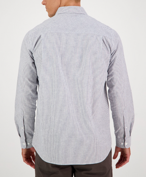 The Swanndri Carillo Beach L/S Shirt, featuring a comfortable blue and white stripe design and long sleeves, is paired with dark pants. The tailored fit enhances the relaxed style as the person stands against a plain white background.