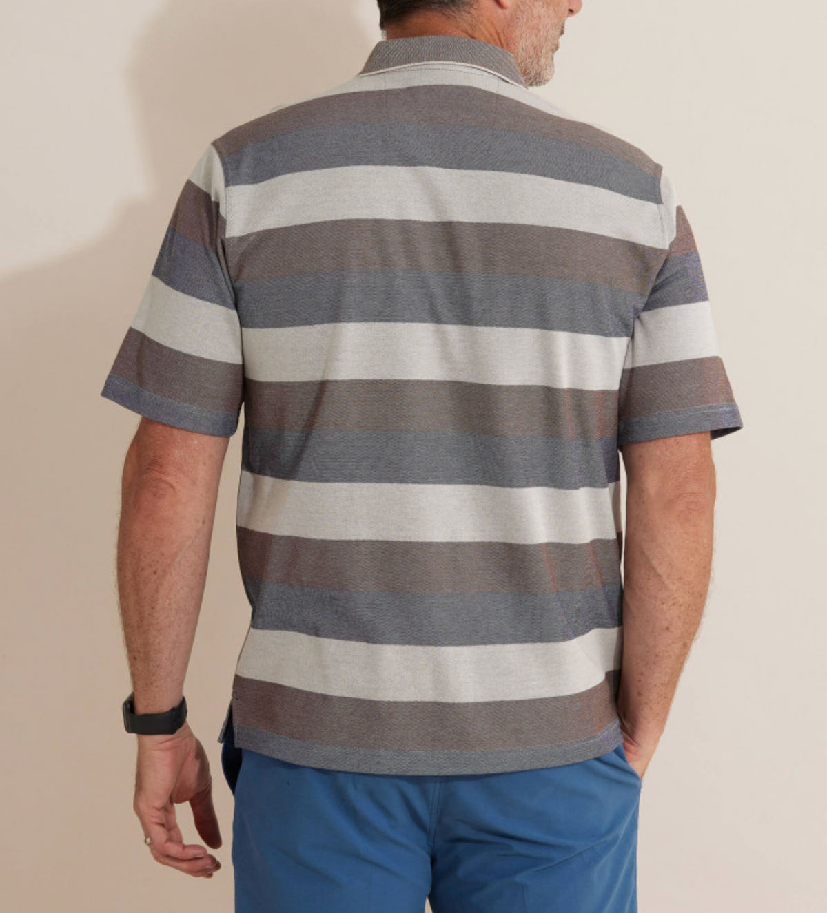 A person with short hair, seen from behind, wears a comfortable Breakaway Machu Micro Prima Polo with stripes in gray and brown and blue shorts. The background is a plain, light-colored wall.
