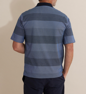 A person wearing the comfortable Breakaway Inva Micro Prima Polo in blue stripes and dark pants stands with their back to the camera. The polo has alternating light and dark blue horizontal stripes on a plain, light-colored background.