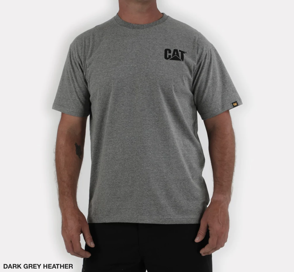 A person dons the casual CAT Trademark Tee in dark grey heather, featuring the classic "CAT" print on the upper left chest. This 100% cotton shirt, paired with black pants, stands out sleekly against a plain white background.