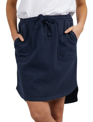 The person is wearing the Foxwood Palm Skirt in navy blue, which features a knee-length design with front pockets and an elastic waistband tied with a drawstring. The skirt's hemline is slightly curved, and it is made from 100% Cotton Baby Cord for extra comfort. They are also wearing a white top and have their hands in the skirt's pockets.