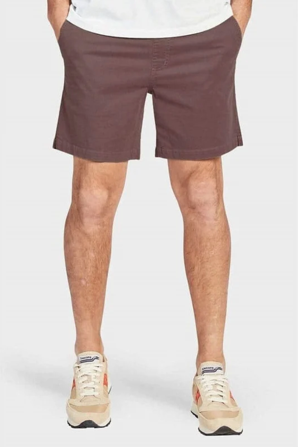A person wearing Academy Brand Volley Shorts in casual brown with an elastic waist and beige sneakers featuring red accents stands with hands in pockets against a white background.