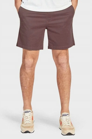 A person wearing Academy Brand Volley Shorts in casual brown with an elastic waist and beige sneakers featuring red accents stands with hands in pockets against a white background.