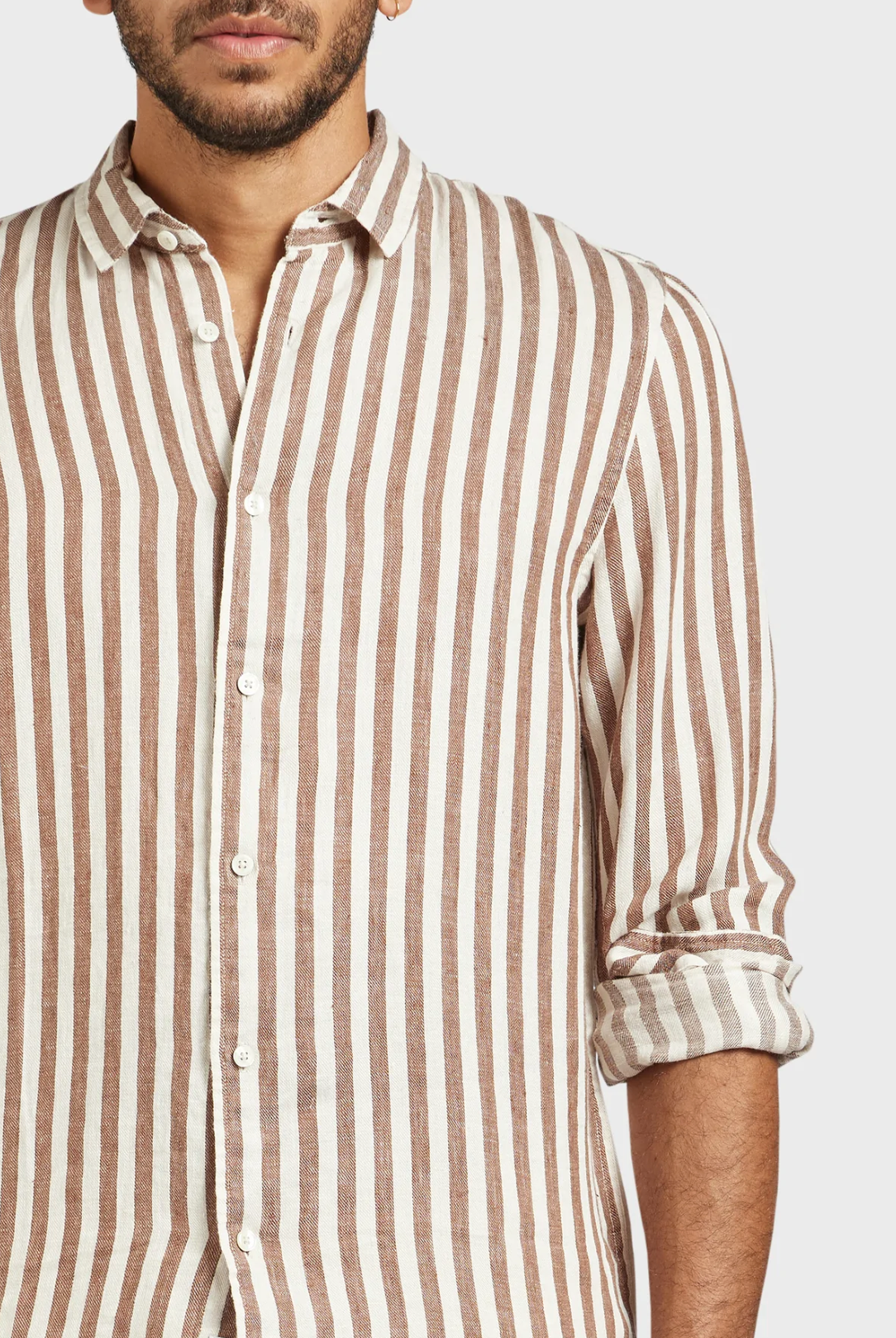 A person is wearing the Academy Brand Farrelly Linen Shirt, featuring long sleeves and brown and white vertical stripes, with the sleeves rolled up to the elbows. The image is cropped to show only their upper body and part of their face against a plain white background. The shirt looks stylish and comfortable.