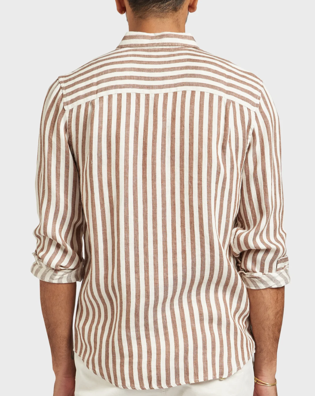 A person is seen from behind wearing a stylish and comfortable Academy Brand Farrelly Linen Shirt, featuring long sleeves, a button-up design, and vertical brown and white stripes. Made of 100% linen, the sleeves are rolled up to the elbows. Part of white pants is visible at the bottom of the image. The background is plain and light-colored.