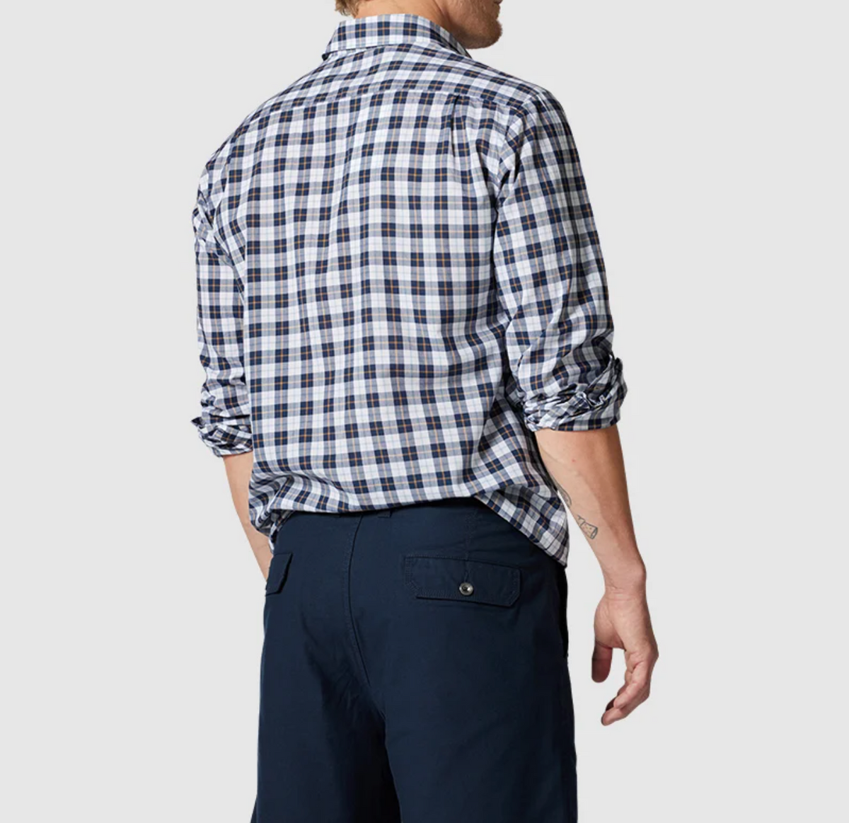 A person with light hair faces away, wearing a Rodd & Gunn Mount Albert Sports Fit Shirt in blue, white, and black plaid. Their slim-fit sleeves are rolled up, paired with dark trousers against a plain gray background.