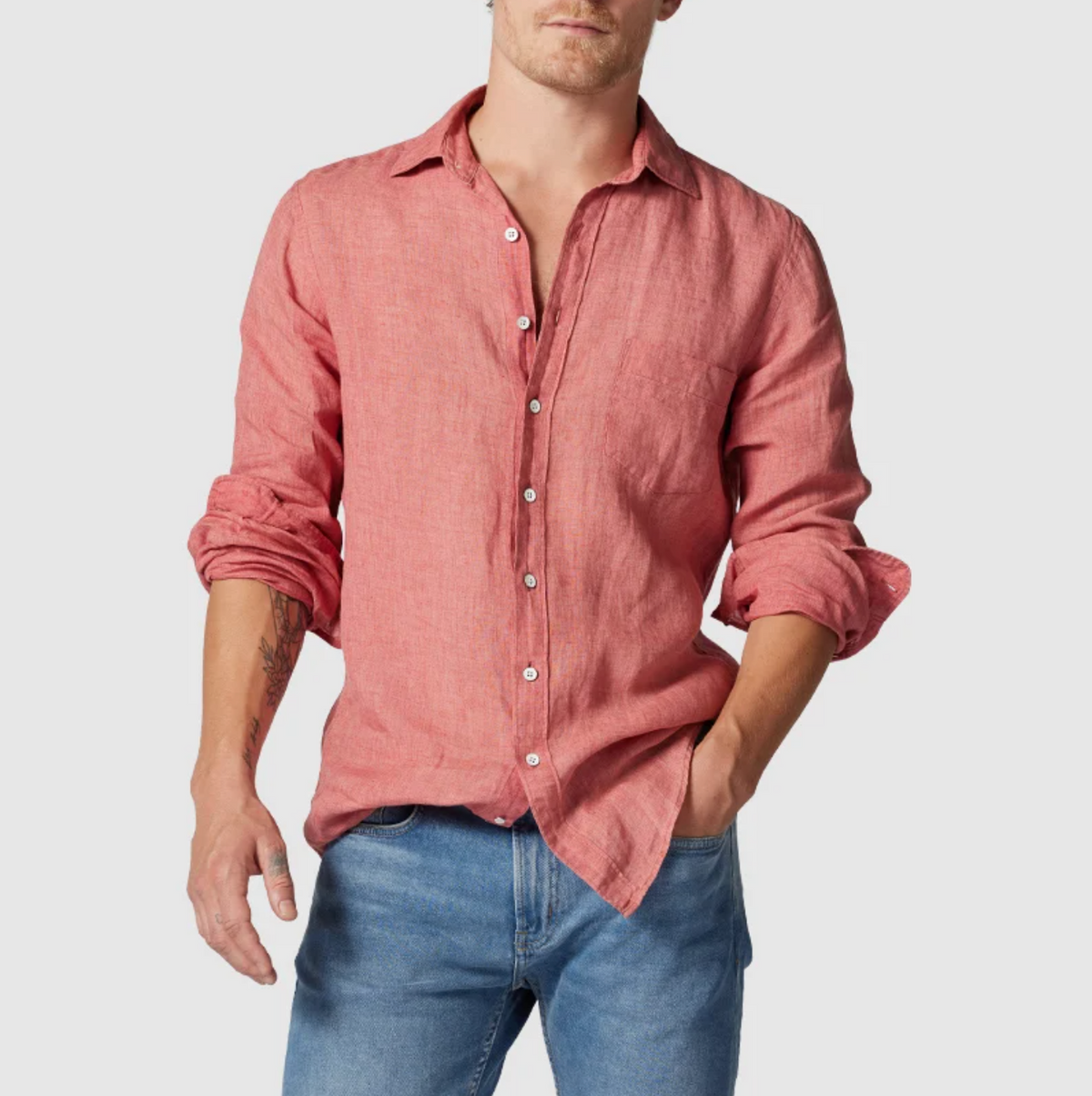 A person in a stylish Rodd & Gunn Coromandel Sports Fit, long-sleeved light red linen shirt with rolled-up sleeves, and blue jeans stands against a plain background, with their right hand in their pocket.