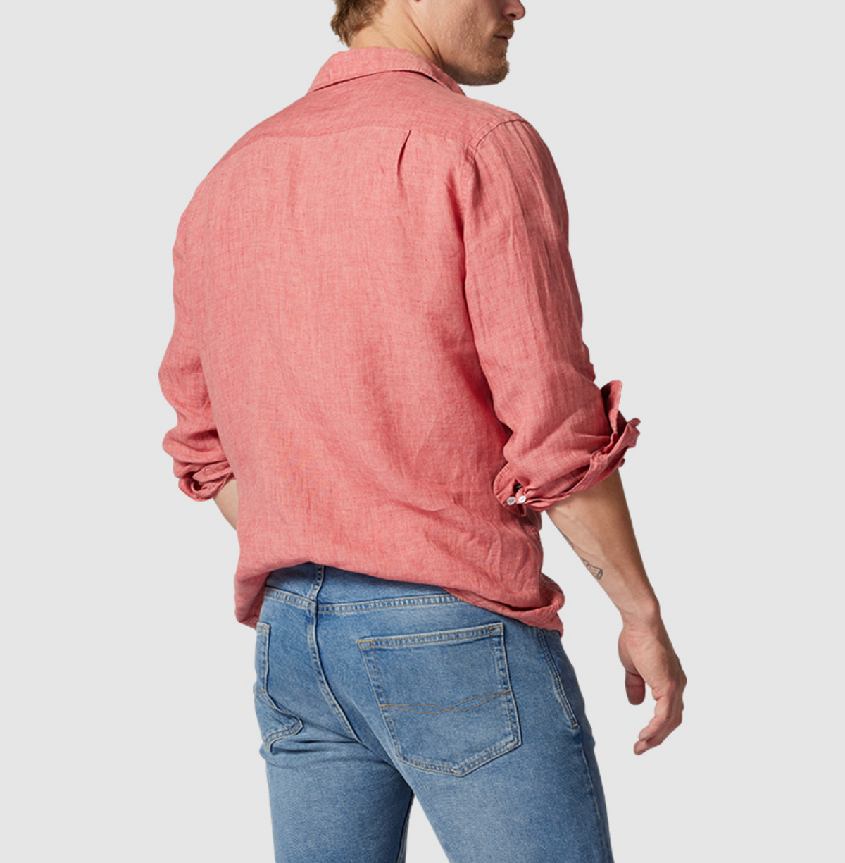 A person faces away from the camera in a Rodd & Gunn Coromandel Sports Fit Shirt, long-sleeved, red, and 100% linen with rolled-up sleeves, paired with slim fit blue jeans against a plain gray backdrop.