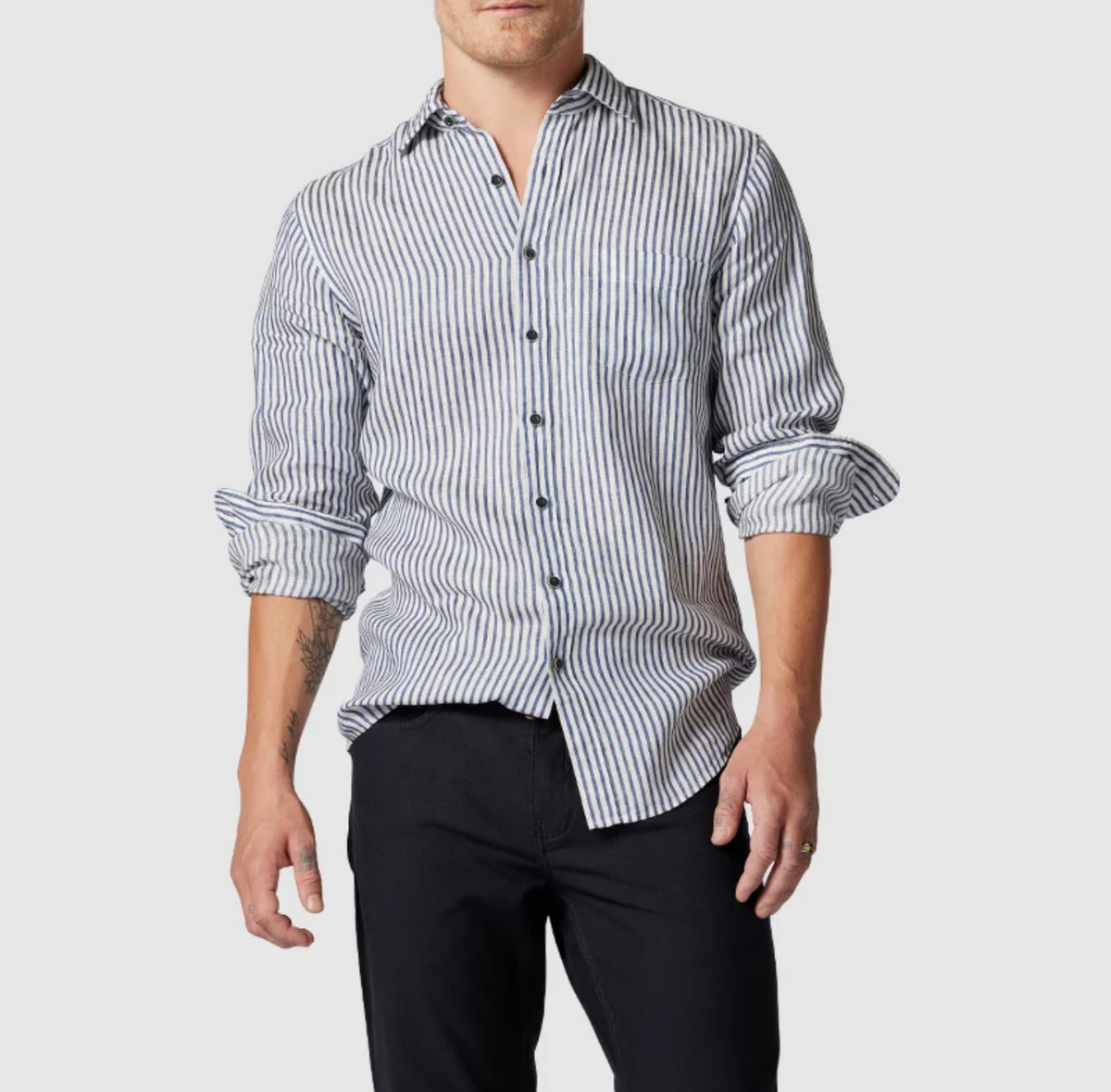 A person wearing a Rodd & Gunn Port Charles Sports Fit Shirt with blue and white stripes crumpled slightly at the sleeves, which are rolled up. The shirt is made from 100% linen, paired with dark pants against a light gray background.