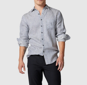 A person wearing a Rodd & Gunn Port Charles Sports Fit Shirt with blue and white stripes crumpled slightly at the sleeves, which are rolled up. The shirt is made from 100% linen, paired with dark pants against a light gray background.
