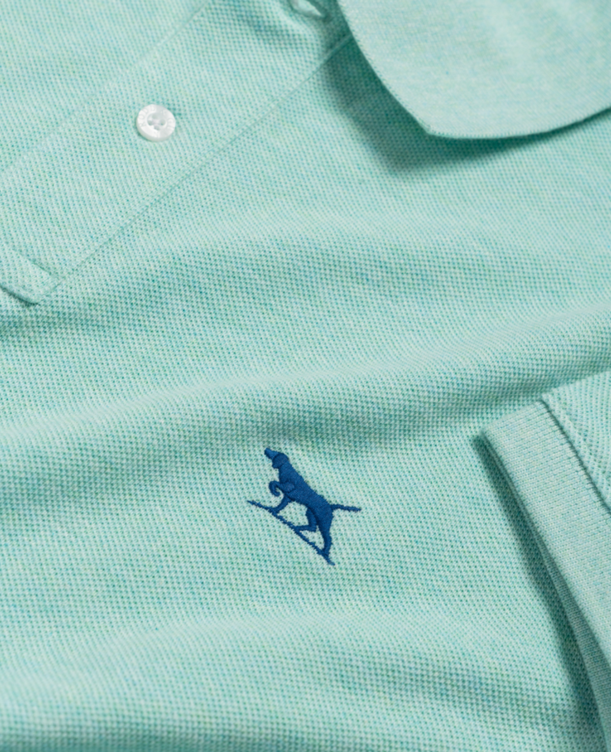 A close-up of the Rodd & Gunn The Gunn Polo in light green, made from premium Australian Cotton, displays a small blue embroidered kangaroo logo near the bottom. This modern-fit polo features textured fabric and a visible white button at the top.