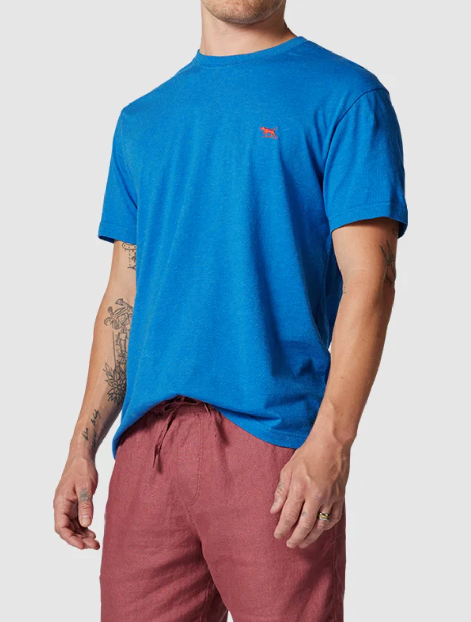 A person displays a sports fit Rodd & Gunn The Gunn T-Shirt, crafted from Australian cotton in blue with a small red chest logo. Paired with red shorts, their tattooed right forearm stands out against a plain backdrop. Their head is unseen as the image crops below the neck.