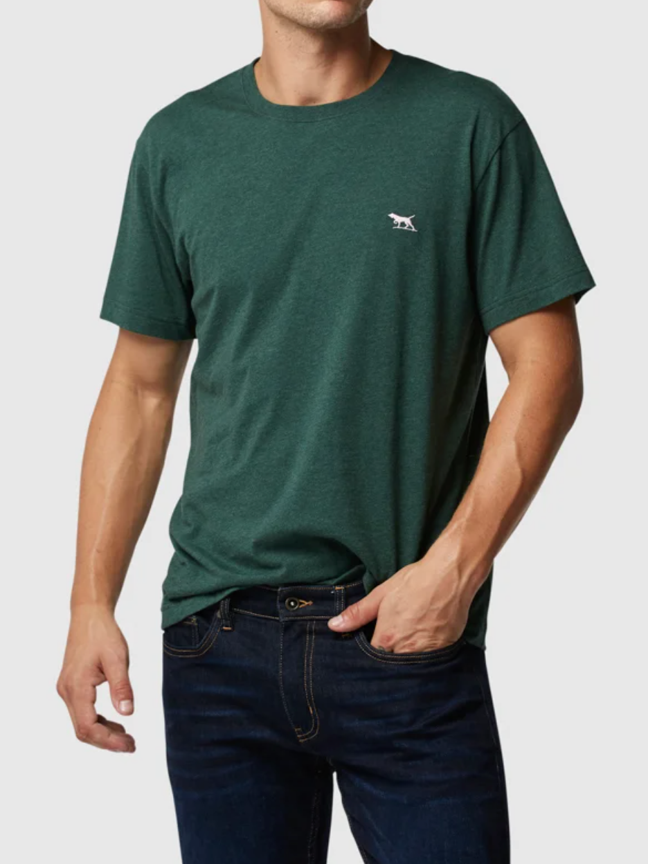 A person is casually standing with one hand in a pocket, wearing the Rodd & Gunn The Gunn T-Shirt in dark green, featuring a small white animal logo on the chest. It's made from Australian cotton and paired with dark blue jeans against a plain gray background.