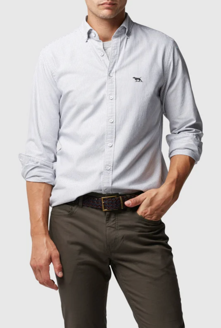 A person wears a Rodd & Gunn Oxford Stripe Sports Fit Shirt, light blue with white stripes, long sleeves partially rolled up against a white background. It's 100% cotton with an embroidered chest logo, paired with dark olive pants and a brown woven belt.