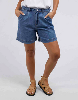 A person wearing a white shirt, Foxwood Melody Shorts made of soft stretch denim, and tan sandals stands with hands in their pockets against a plain background.